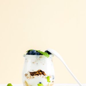 Yogurt oat granola with jam, blueberries and green leaves in glass jar on pastel yellow backdrop.